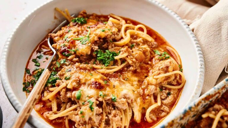 A bowl of spaghetti topped with meat sauce, melted cheese, and chopped parsley, with a fork resting inside.
