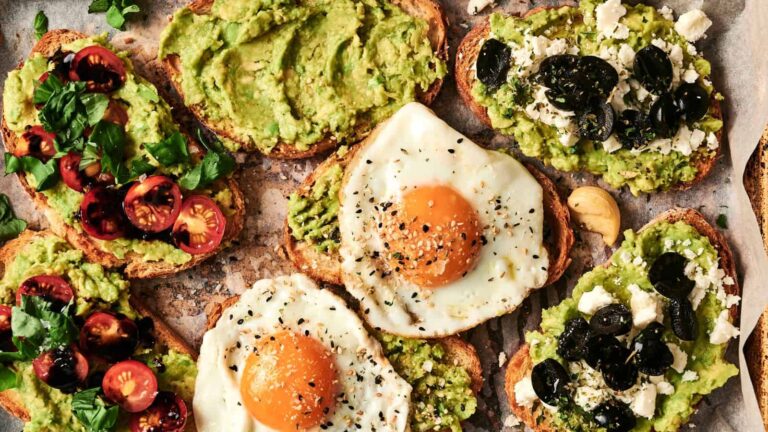 Various avocado toasts with toppings including cherry tomatoes, fried eggs, black olives, and feta cheese on a parchment-lined surface.