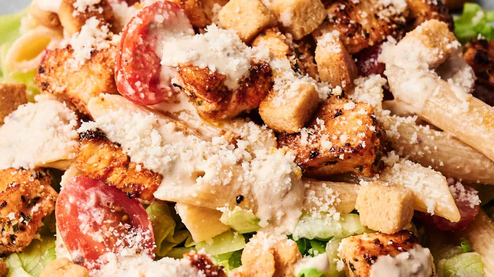 A plate of pasta salad with grilled chicken pieces, romaine lettuce, croutons, and creamy dressing.