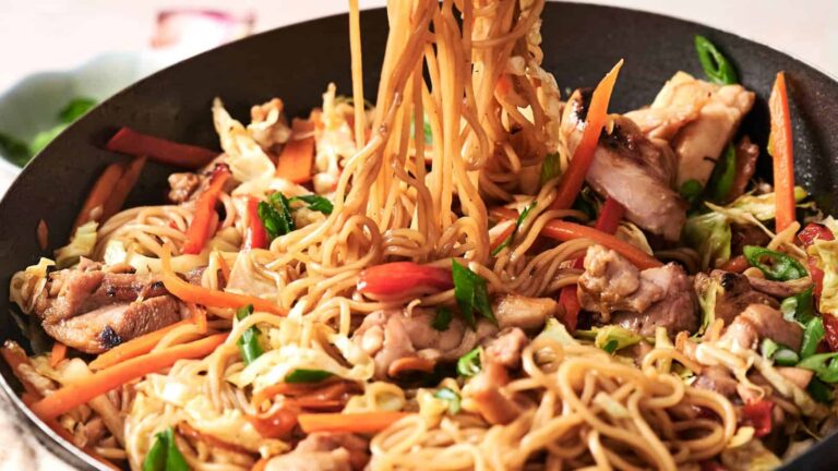 Stir-fried noodles with vegetables and chicken in a pan, featuring carrots, cabbage, and green onions.