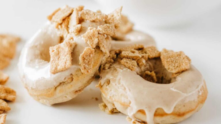 Two glazed donuts topped with crumbled cereal pieces are placed on a white surface.