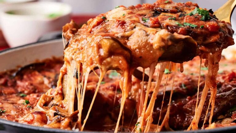 A slice of cheesy eggplant lasagna with tomato sauce is being lifted from a pan, showing stretchy melted cheese. A bowl is blurred in the background.