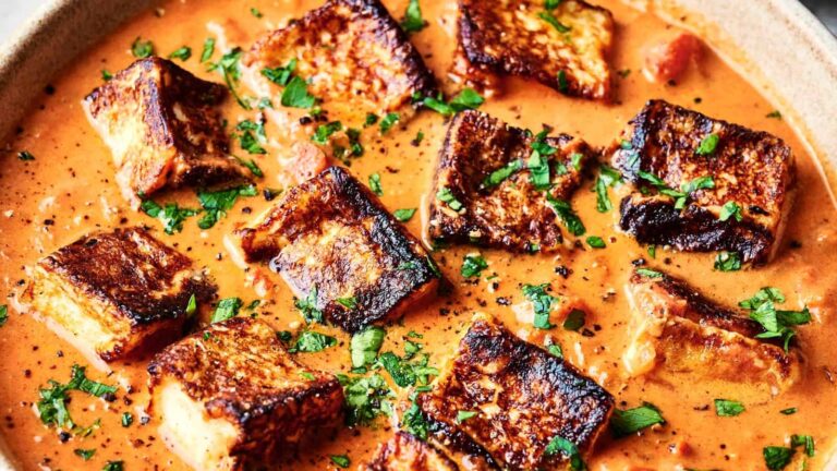 A dish of grilled paneer cubes in a rich, orange curry sauce, garnished with chopped cilantro, served in a shallow bowl.