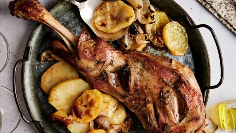 Roasted lamb leg on a platter with sliced potatoes, lemon, and herbs. A knife is placed beside the dish.