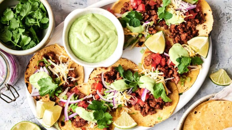 A plate of beef tacos topped with cheese, tomatoes, red onions, and cilantro, served with avocado sauce and lime wedges. A bowl of cilantro and a jar lid are next to the plate.