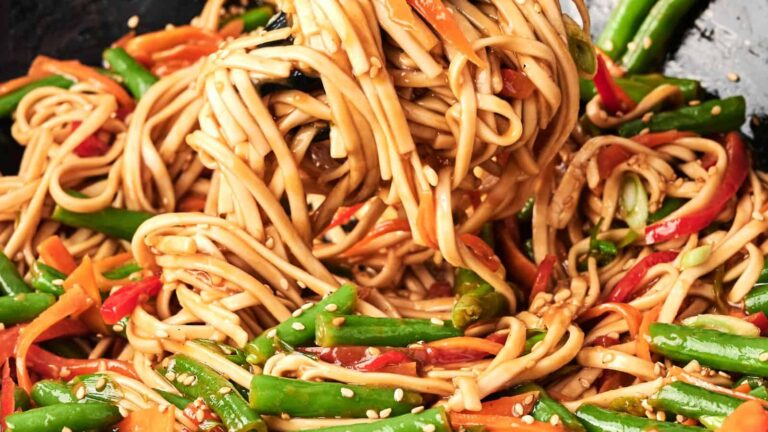 A close-up of stir-fried noodles with green beans, red bell peppers, and carrots, garnished with sesame seeds.