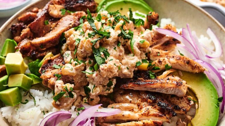 A bowl of rice topped with grilled chicken, creamy sauce, sliced avocado, chopped cilantro, and red onion slices.
