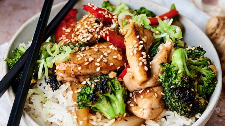 A bowl of rice topped with broccoli, chicken, red peppers, green onions, and sesame seeds, with black chopsticks resting on the side.