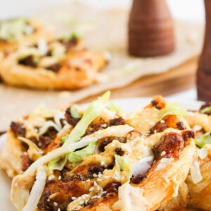 Close-up of Big Mac Pinwheels featuring puff pastry topped with ground meat, shredded lettuce, sesame seeds, and a drizzle of creamy sauce on a white plate, with more pastries and pepper mills in the background.