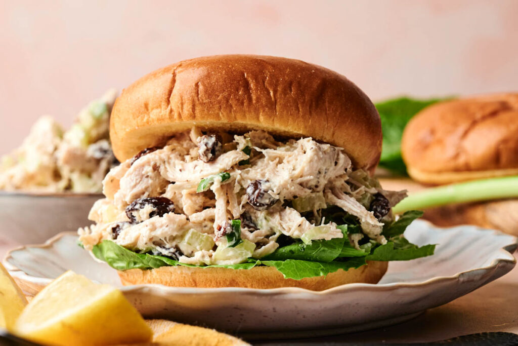 Chicken Salad Sandwich with lettuce on a bun, served on a plate. This classic sandwich features shredded chicken, raisins, and celery in the filling for a delicious mix of flavors.
