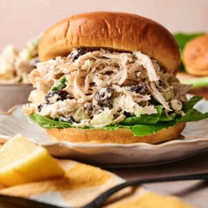 A Chicken Salad Sandwich with lettuce and dark raisins on a bun, served on a plate with a lemon wedge nearby.