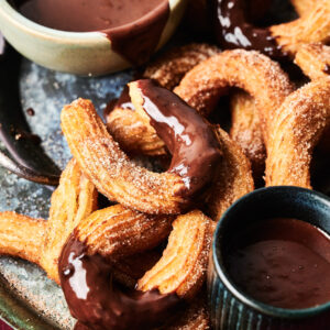 A plate of churros with dipping sauce, coated in cinnamon sugar, some dipped in chocolate sauce, with two bowls of chocolate sauce on the side.