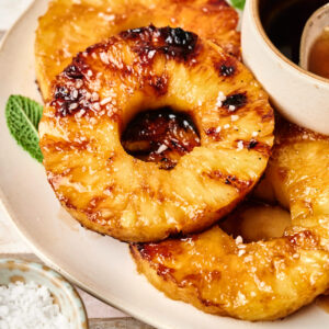 Grilled Pineapple rings on a plate, garnished with coarse salt and mint, are served with a small bowl of sauce nearby.