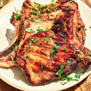 Two Grilled Pork Chops, bone-in and garnished with chopped fresh parsley, are served on a white plate.