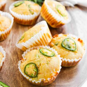 Jalape&ntilde;o Honey Cornbread Muffins are topped with sliced jalape&ntilde;os and arranged on a wooden board, accented by fresh herbs and a sliced jalape&ntilde;o in the background.