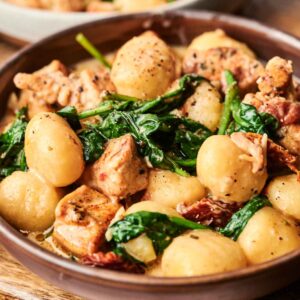A bowl of Marry Me Chicken Gnocchi with spinach and sun-dried tomatoes in a creamy sauce, served on a wooden board.