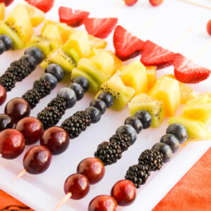 Rainbow Fruit Skewers featuring grapes, blackberries, blueberries, kiwi, pineapple, and strawberries are arranged on a white surface, with extra fruit and a red cloth nearby.