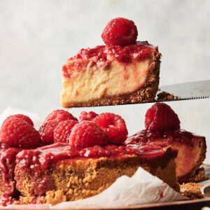 A slice of Raspberry Cheesecake is being lifted from a whole cheesecake topped with raspberry sauce and fresh raspberries, displayed on a wooden cake stand.