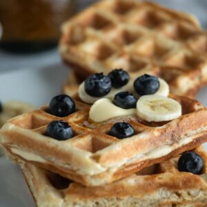 Two stacked sourdough waffles topped with sliced bananas, blueberries, and butter on a white plate.