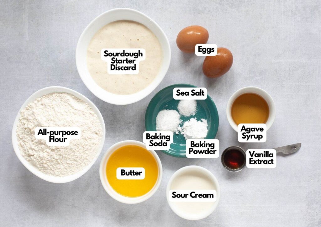 A top-down view of bowls containing ingredients for Sourdough Waffles: sourdough starter discard, flour, butter, sour cream, sea salt, baking soda, baking powder, agave syrup, vanilla extract, and two eggs.