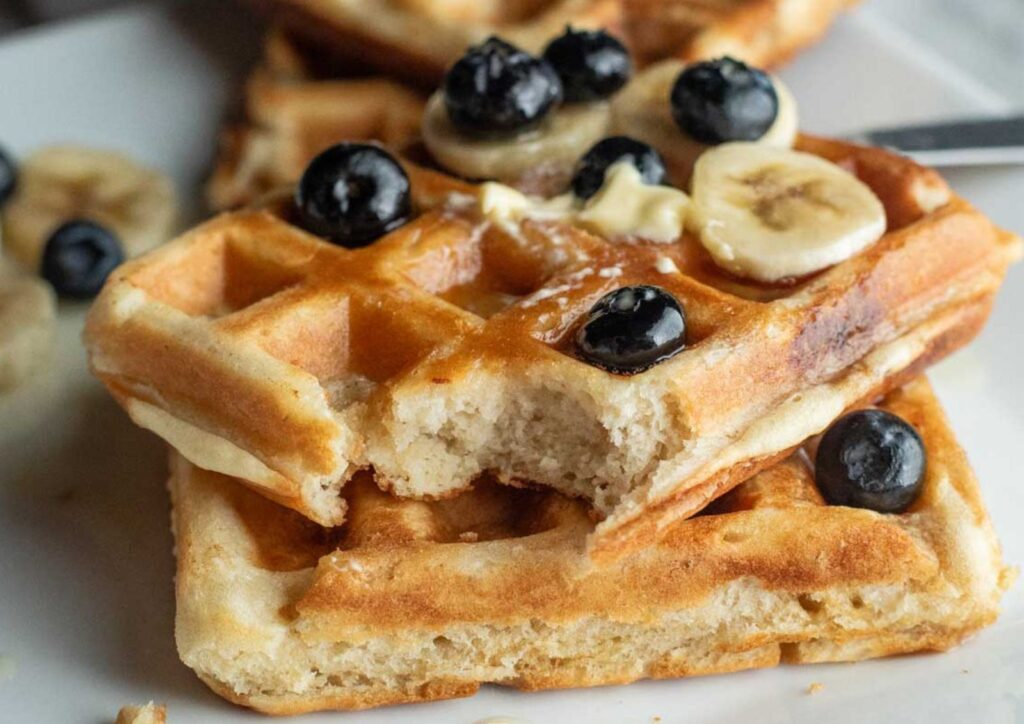 A stack of sourdough waffles with a bite taken out, topped with banana slices, blueberries, butter, and syrup on a white plate.