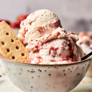 A bowl of Strawberry Shortcake Ice Cream swirled with strawberries, topped with a piece of shortbread cookie and garnished with a fresh strawberry.