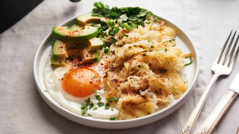 A breakfast plate with a sunny-side-up egg, sliced avocado, hash browns, and fresh herbs, served with a fork and knife on a white tablecloth.