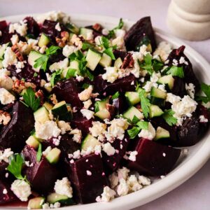 A plate of Roasted Beet Salad topped with chopped cucumber, crumbled feta cheese, parsley, and pecans on a light surface.