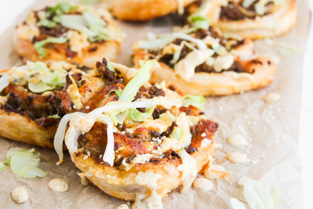 Big Mac Pinwheels: Mini pizzas topped with ground beef, shredded lettuce, sesame seeds, and creamy sauce on parchment paper.