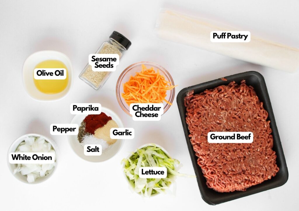 Top-down view of ingredients for Big Mac Pinwheels: ground beef, cheddar cheese, puff pastry, olive oil, sesame seeds, paprika, pepper, salt, garlic, white onion, and lettuce.