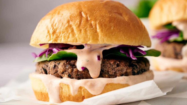 A close-up of a veggie burger with leafy greens, shredded purple cabbage, and creamy sauce on a brioche bun.