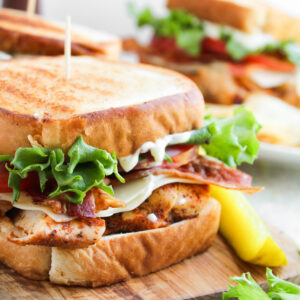 A toasted Blackened Chicken Club Sandwich with grilled chicken, bacon, lettuce, tomato, and cheese on a wooden board, garnished with a pickle spear and fresh lettuce.