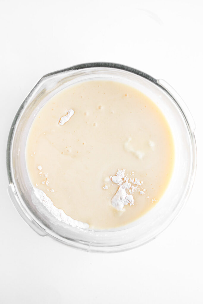 A glass mixing bowl containing a pale batter with a few lumps of flour on the surface, viewed from above on a white background.