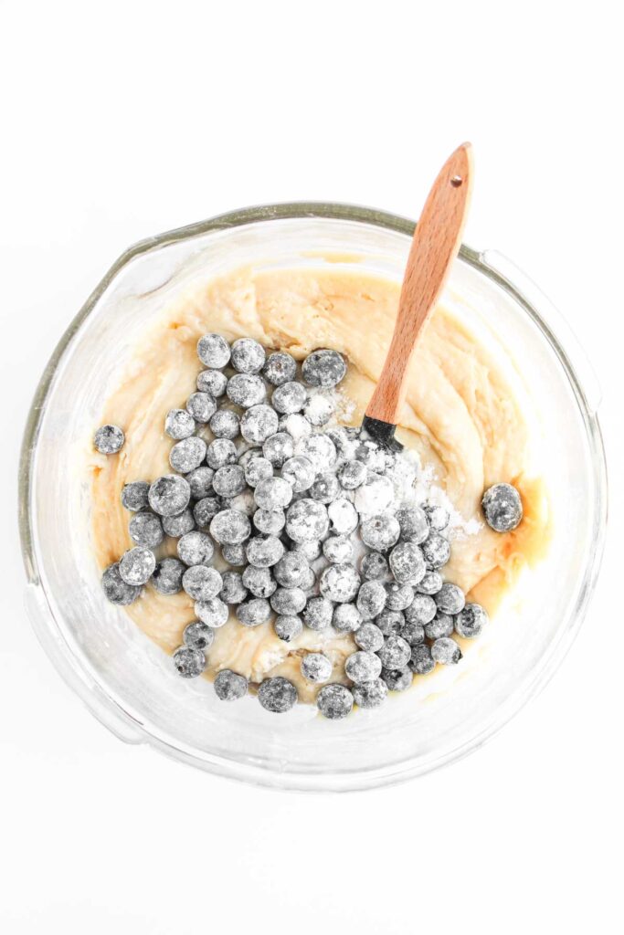 A glass bowl containing batter with a pile of blueberries and a wooden spatula, ready to be mixed.
