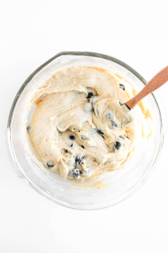 A glass bowl containing thick batter with blueberries being mixed with a wooden spoon, viewed from above on a white background.