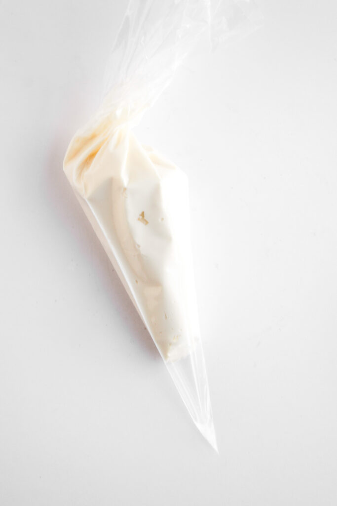 A clear plastic piping bag filled with white cream or frosting is placed on a white surface.