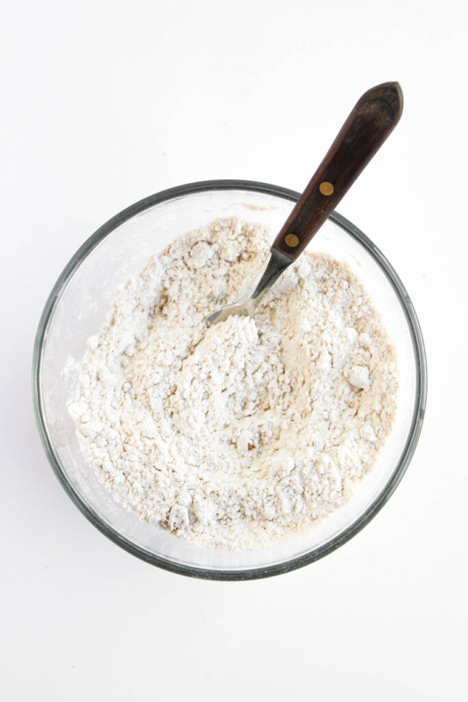 A glass bowl filled with flour mixture and a metal fork with a dark handle, set on a white background.