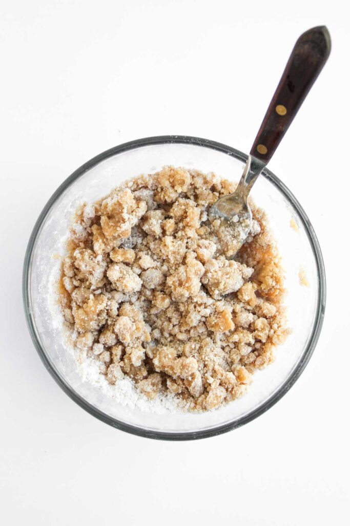 A glass bowl filled with a crumbly brown sugar and flour mixture, partially mixed with a fork, on a white background.