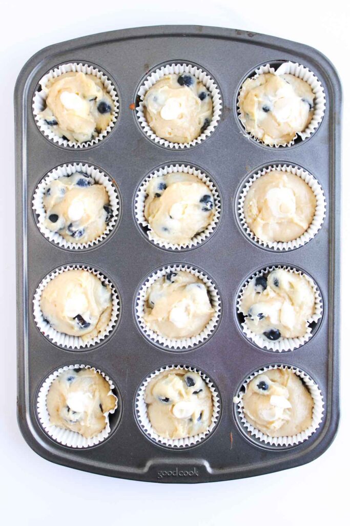 A muffin tin with twelve paper liners filled with unbaked blueberry muffin batter.