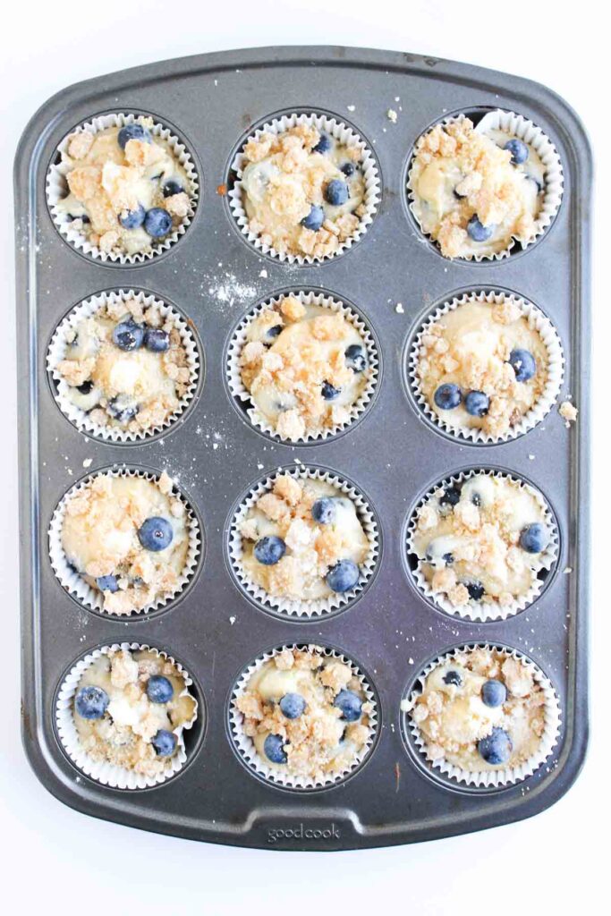 A muffin tin filled with twelve unbaked blueberry muffins in paper liners, topped with streusel and fresh blueberries.