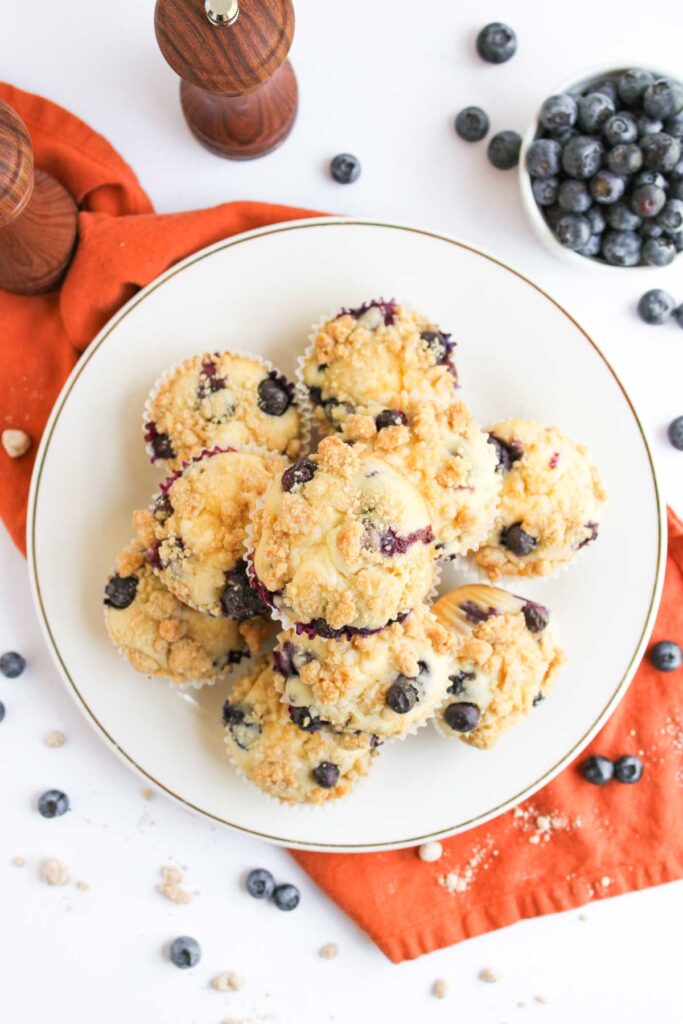 A plate of blueberry muffins with crumb topping sits on an orange napkin, surrounded by scattered fresh blueberries and wooden pepper mills.
