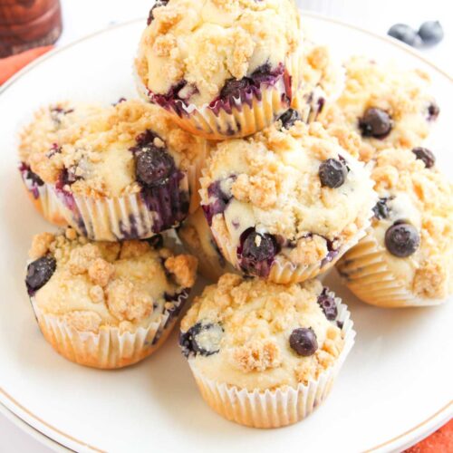 A plate of blueberry muffins with crumb topping is arranged on a white plate, with fresh blueberries in a bowl in the background.