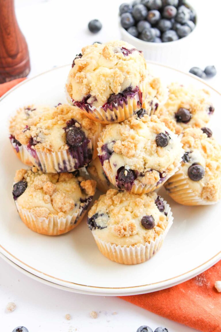 A plate of blueberry muffins with crumb topping is arranged on a white plate, with fresh blueberries in a bowl in the background.