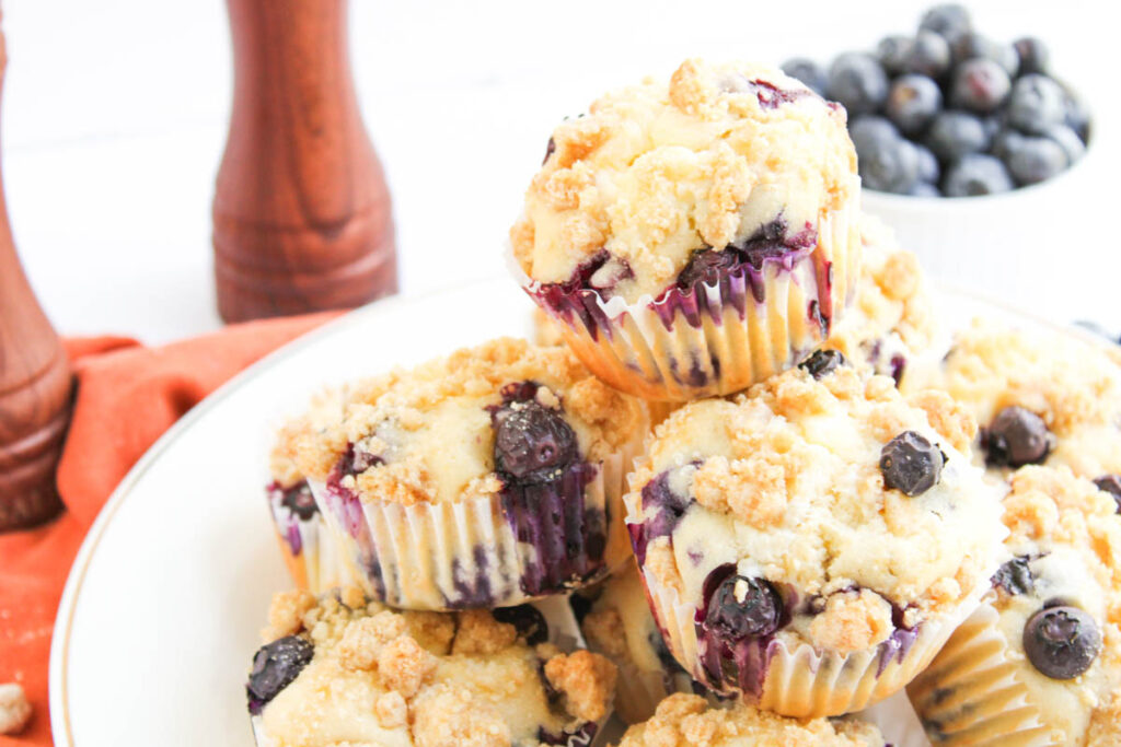 A plate of blueberry muffins with crumb topping sits on a white surface; a bowl of fresh blueberries is in the background.