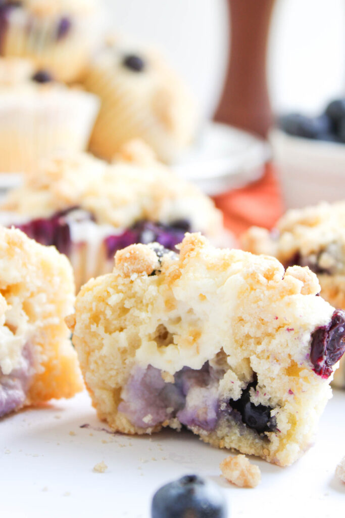 A close-up of a blueberry muffin cut in half, showing a crumbly topping and visible blueberries inside, with whole muffins and blueberries in the background.