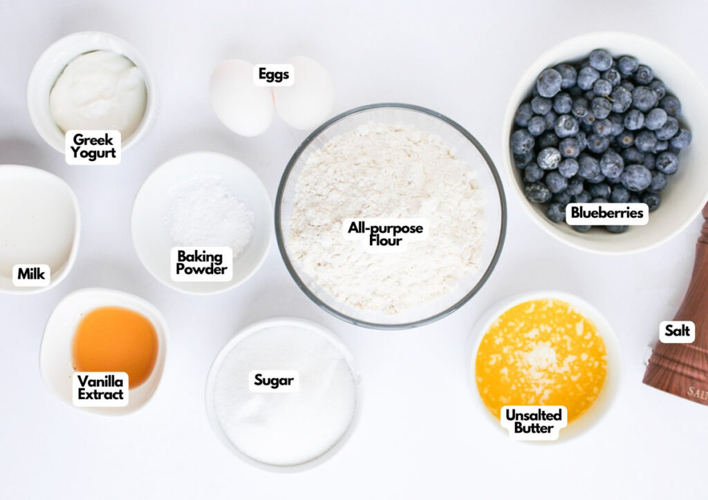 Top-down view of labeled baking ingredients in bowls: Greek yogurt, milk, vanilla extract, eggs, baking powder, sugar, all-purpose flour, blueberries, unsalted butter, and salt.