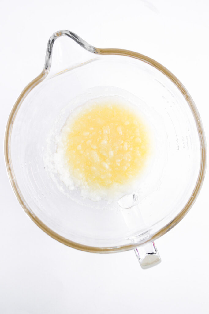 A glass mixing bowl containing partially mixed melted butter and sugar on a white background.