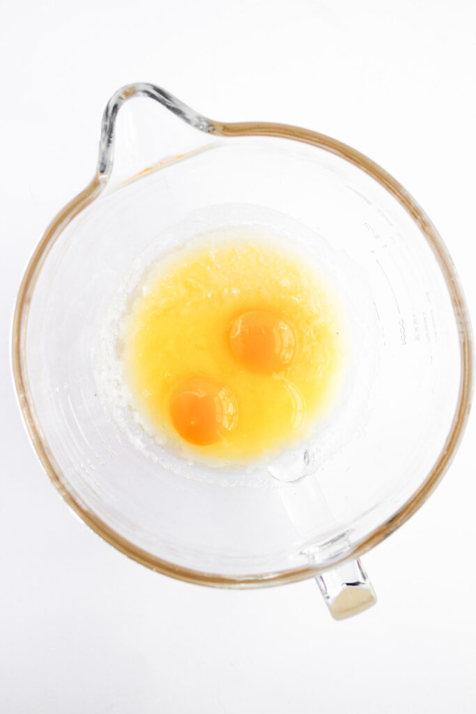 Two raw eggs cracked into a glass mixing bowl with some liquid, viewed from above on a white background.