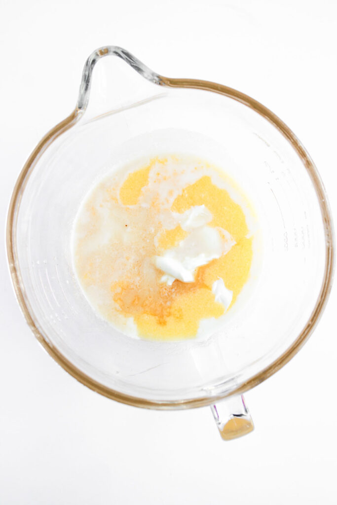 A glass mixing bowl containing eggs, milk, melted butter, and a dollop of yogurt or sour cream, viewed from above on a white background.