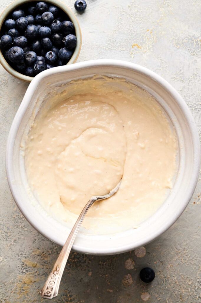 A bowl of pancake batter with a spoon inside is next to a small bowl of fresh blueberries on a light surface.